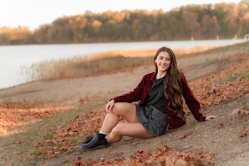 A person sits on autumn ground by a lake wearing a burgundy cardigan and gray dress with boots at sunset.