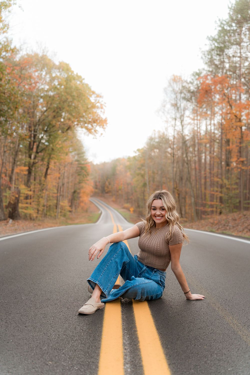 A person in jeans and sweater sits on yellow lines of autumn forest road with colorful fall foliage.