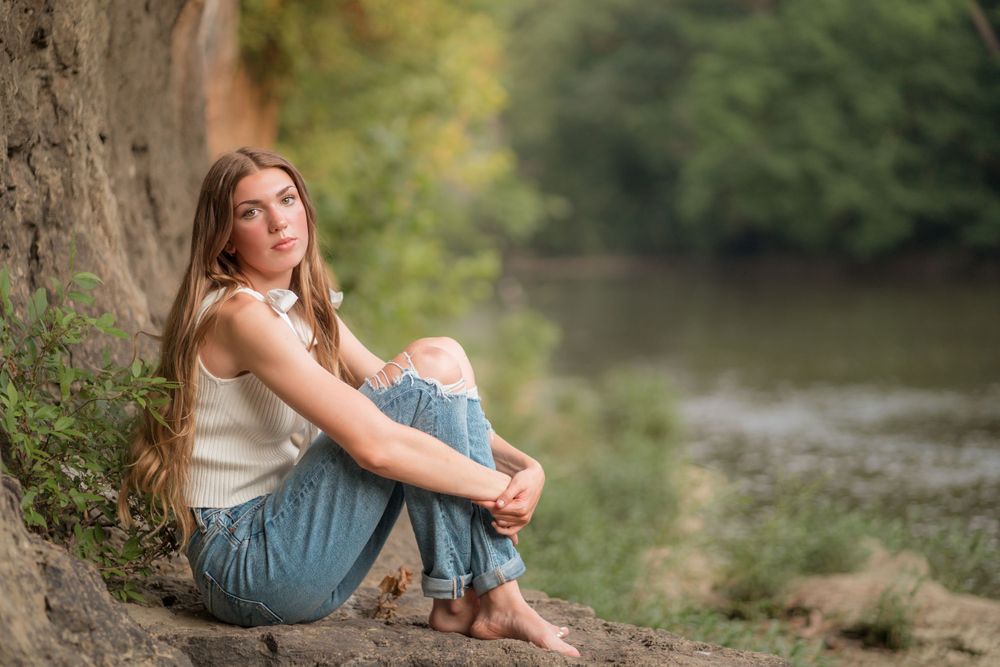 A person in a cream sweater and jeans sits thoughtfully by a riverbank under a tree on an autumn afternoon.
