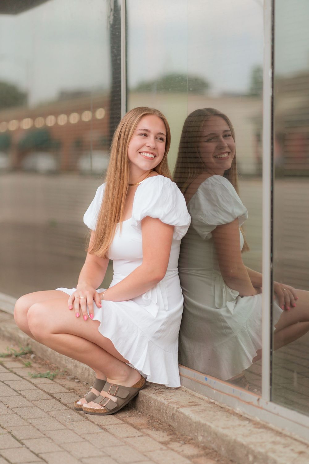 Person in a white summer dress crouches by a reflective window while posing for a photo outdoors.