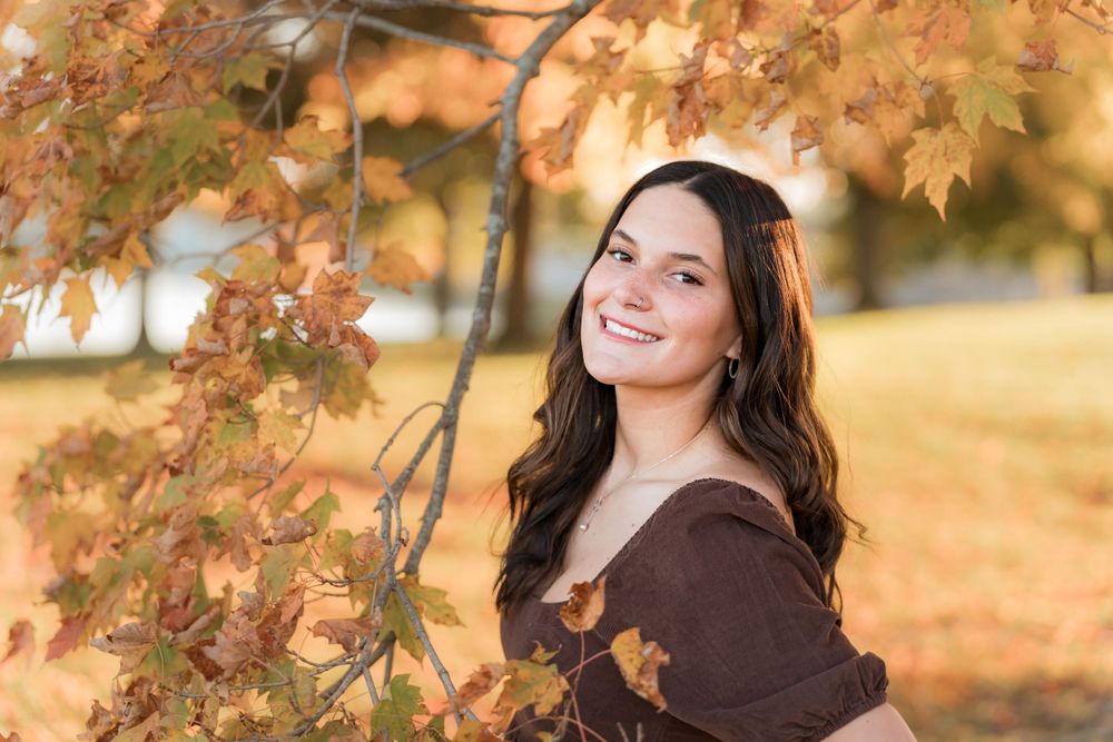 A person poses next to autumn maple leaves in warm sunlight wearing a brown sweater outdoors during golden hour.