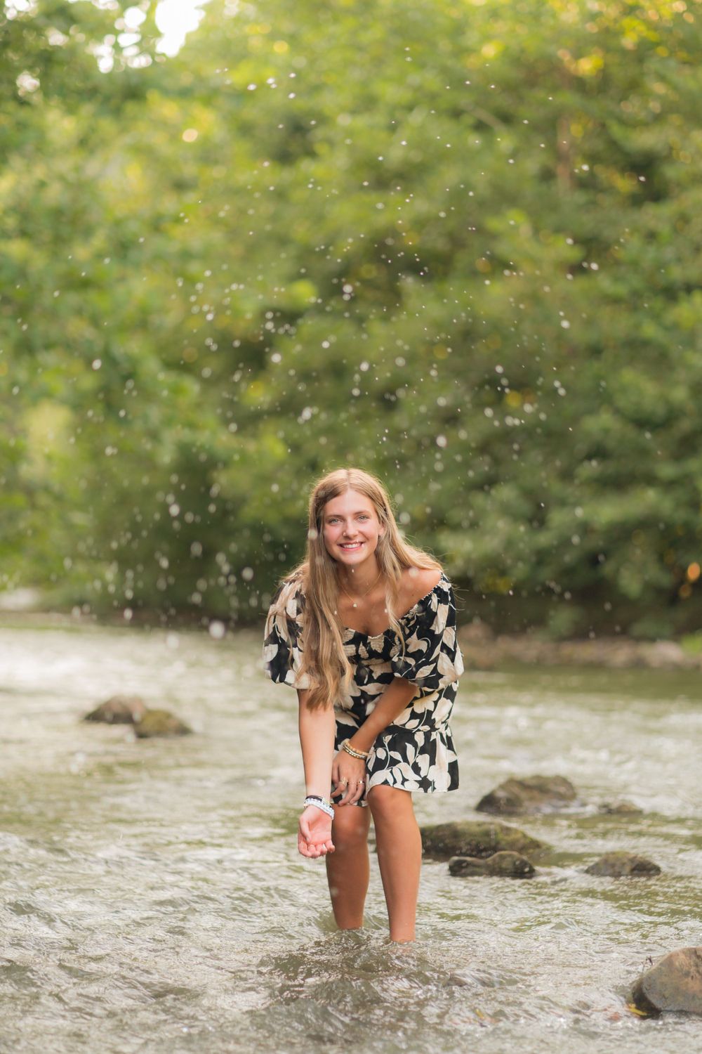A floral dress-clad figure splashes playfully in a rocky stream while water droplets sparkle in the sunlight.