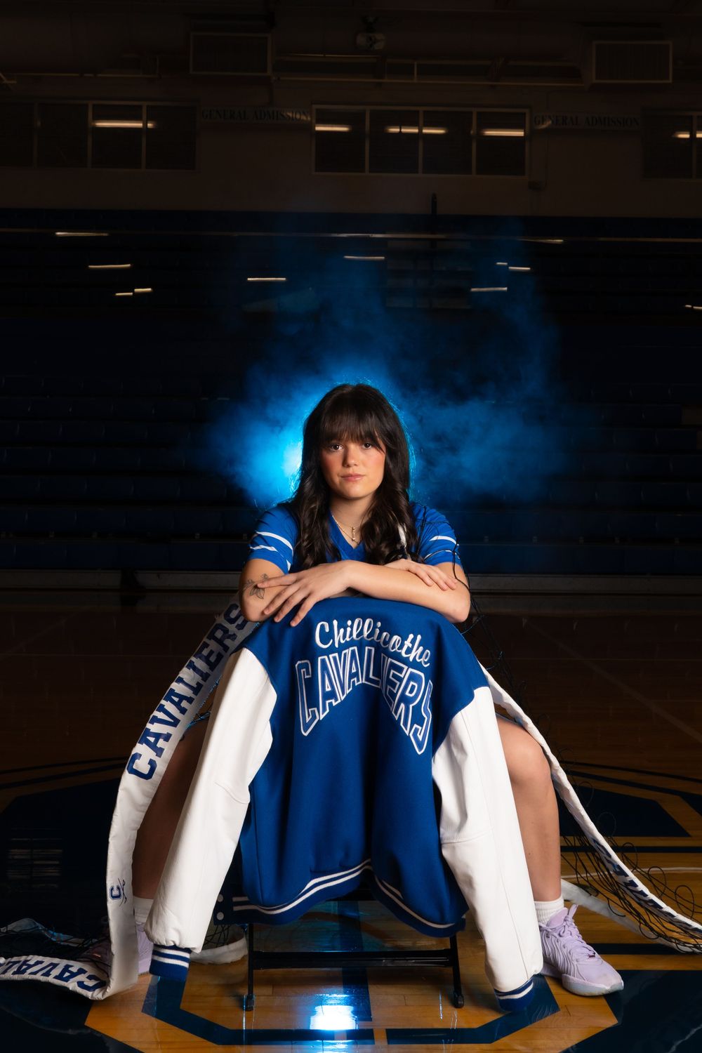 Student athlete sitting in a chair on a basketball court wearing a blue and white varsity jacket with dramatic lighting.