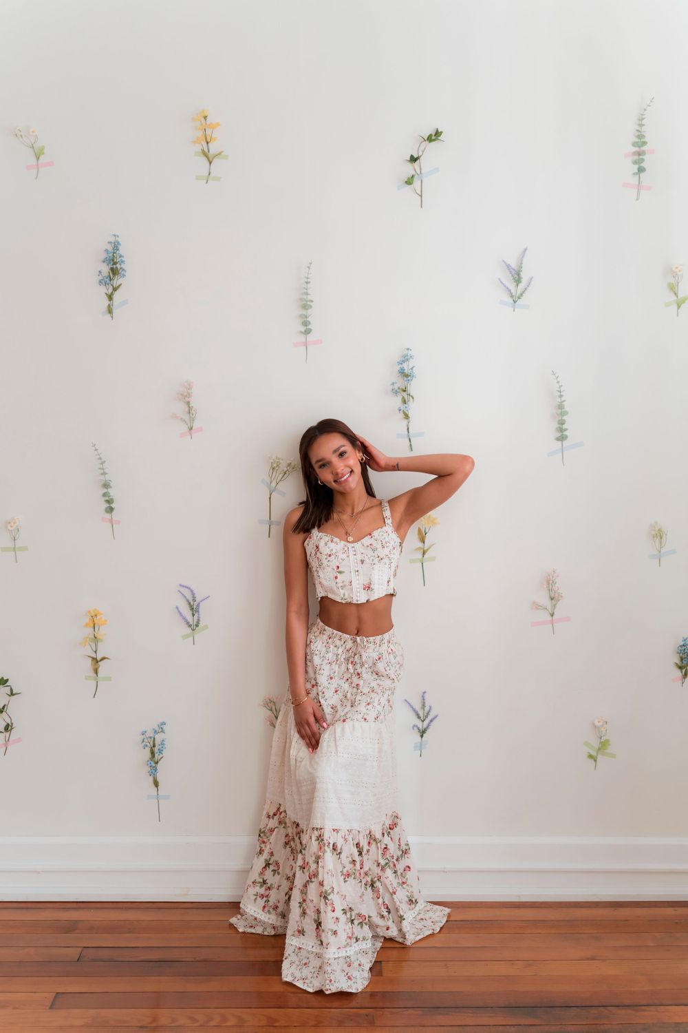 A model in a two-piece white lace dress poses against a floral decorated wall with wooden flooring.