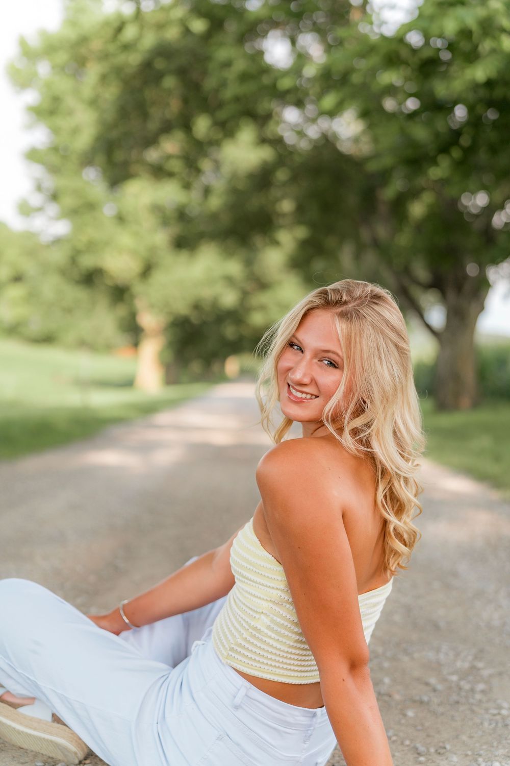 A woman in a white dress lies on a gravel path surrounded by green trees in a summer outdoor setting.