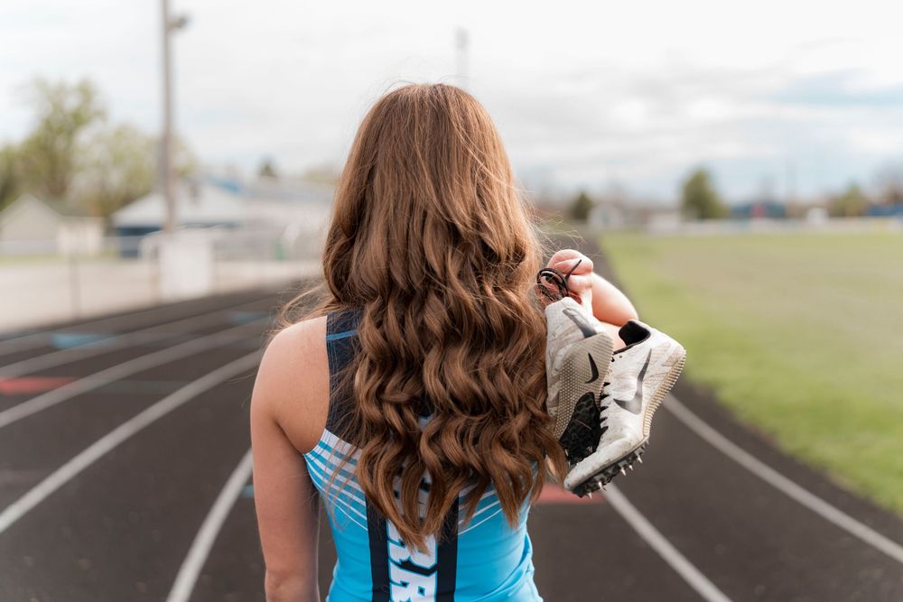 Athletic woman with long wavy brown hair standing on a running track wearing blue workout attire.