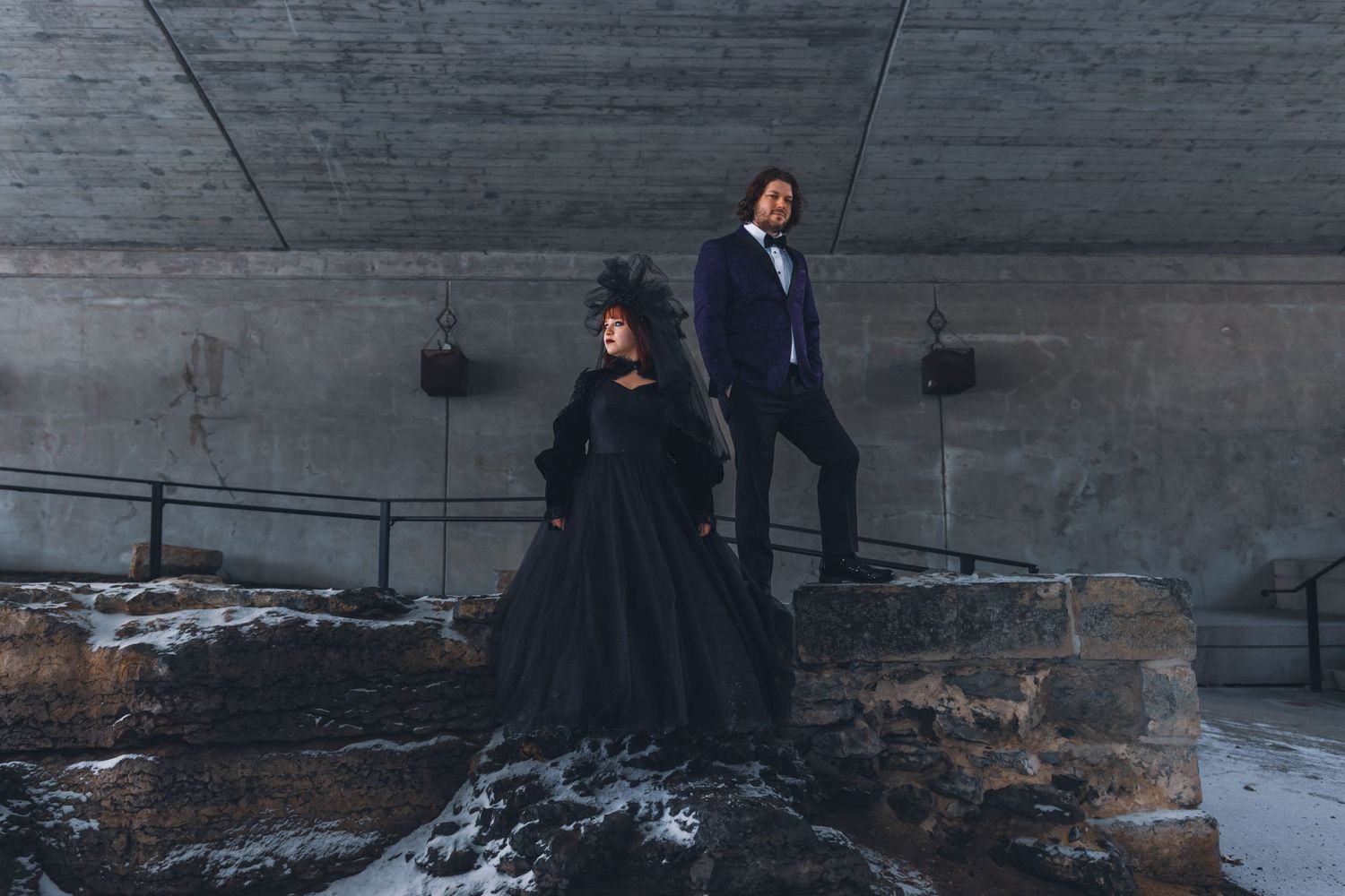 Two people in elegant black attire pose dramatically on snowy concrete steps against a gray wall.