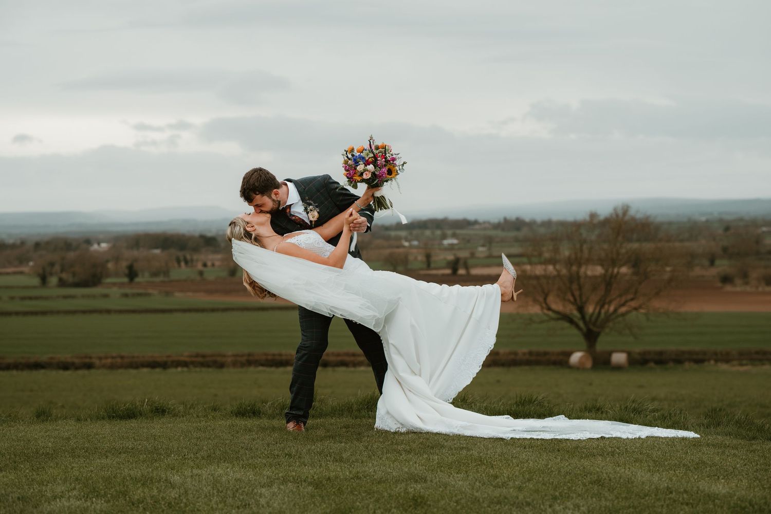Romantic dip pose wedding photo with flowing dress against countryside landscape.