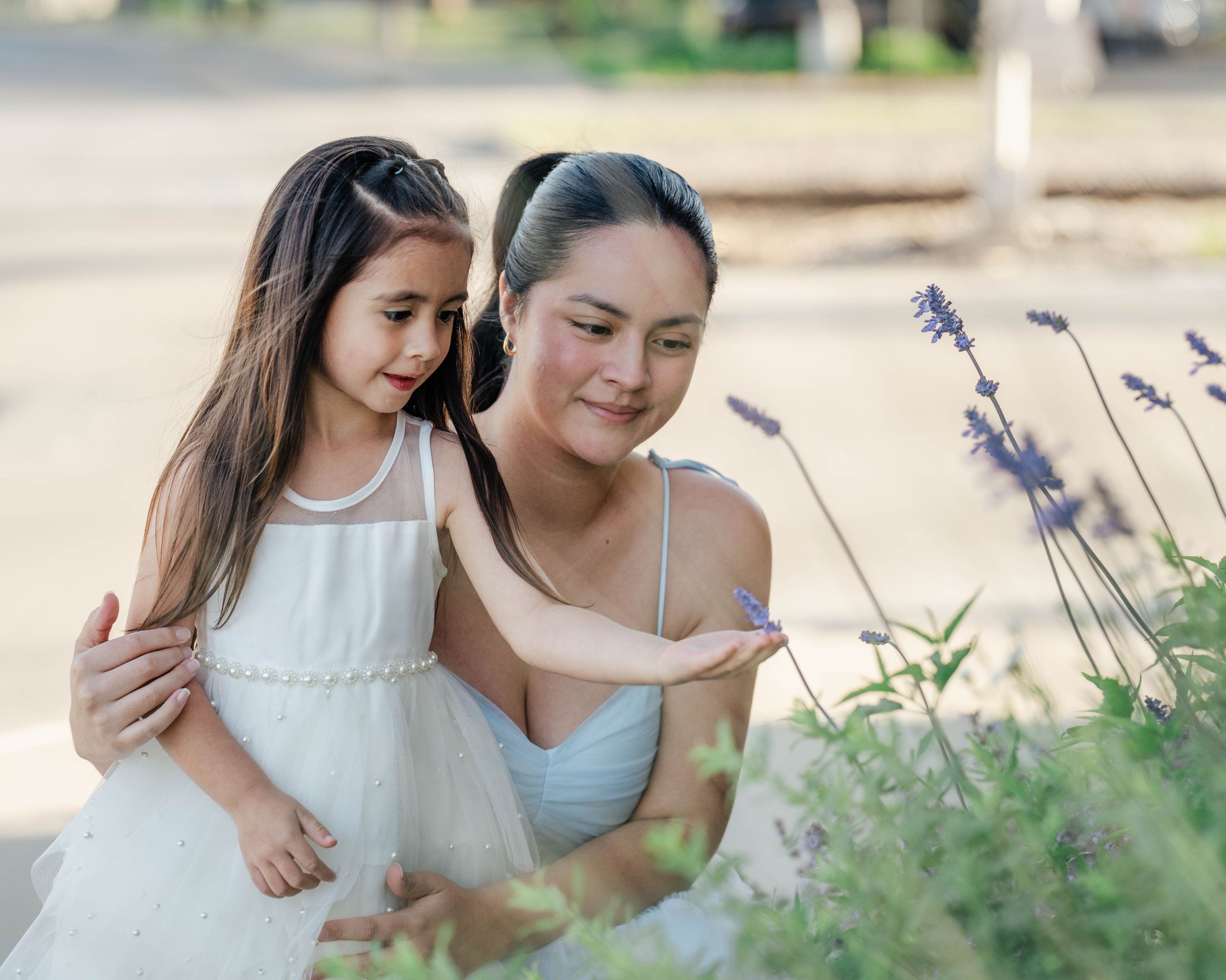 Parent and child in white dresses examine purple flowers growing in a garden.