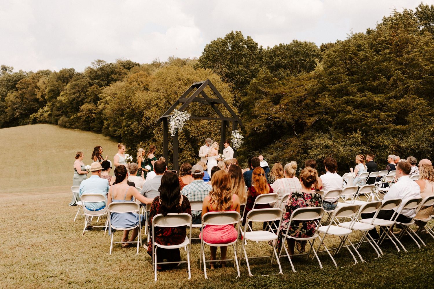 Outdoor woodland wedding ceremony with couple at floral arch, bridesmaids in blue, and guests seated on white chairs.
