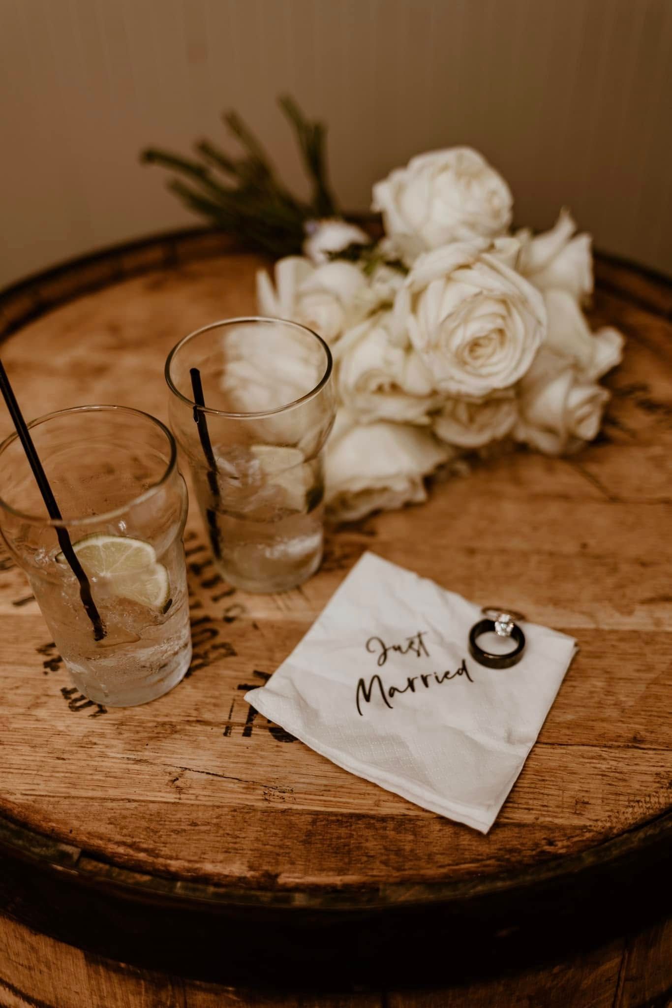 White roses and empty cocktail glasses sit on a rustic wooden barrel top with a handwritten 'Just Married' napkin.