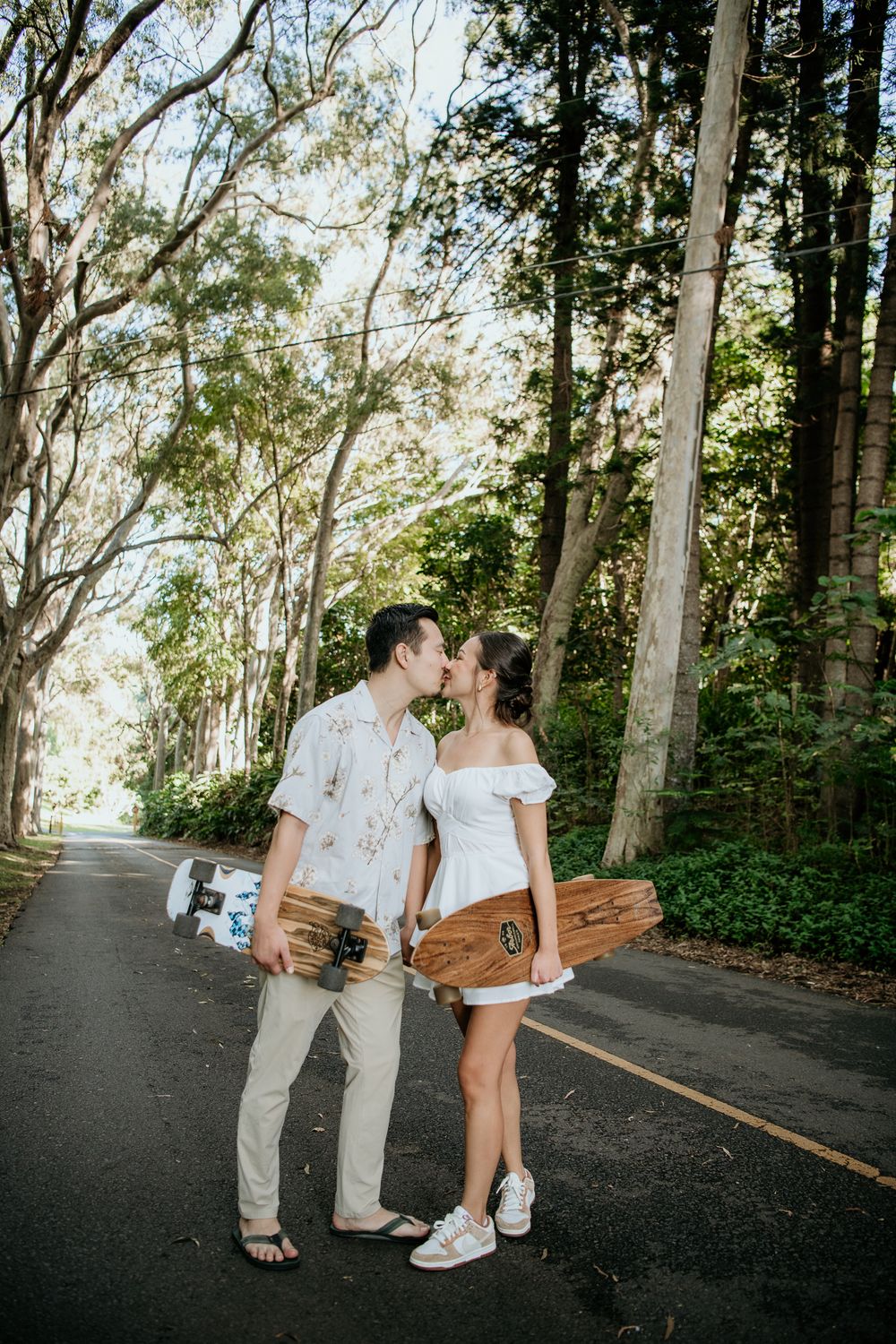 A couple walking and sharing an intimate moment on a tree-lined road while carrying skateboards.
