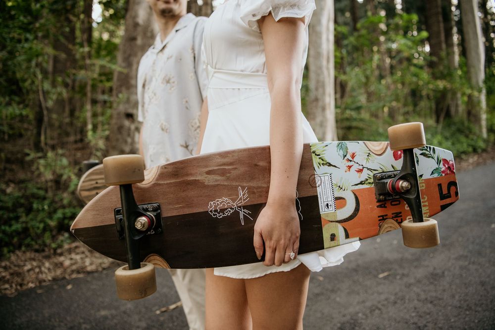 Person in a white dress holding a brown longboard with colorful abstract designs along a wooded path.