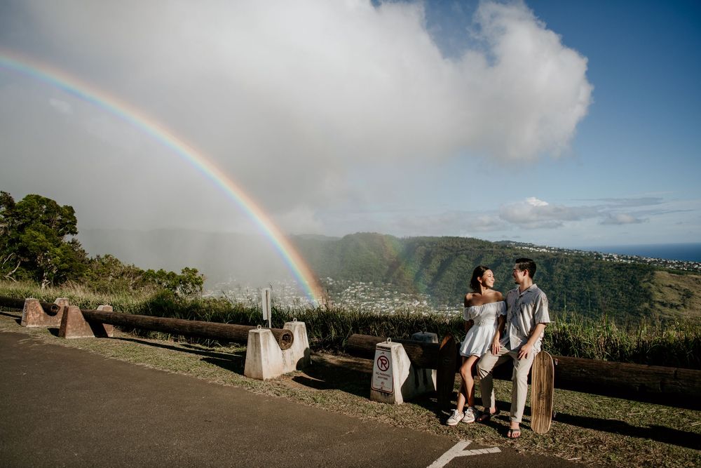 Scenic mountain overlook with a vibrant rainbow arching across the sky while two people enjoy the view from a roadside rest area.