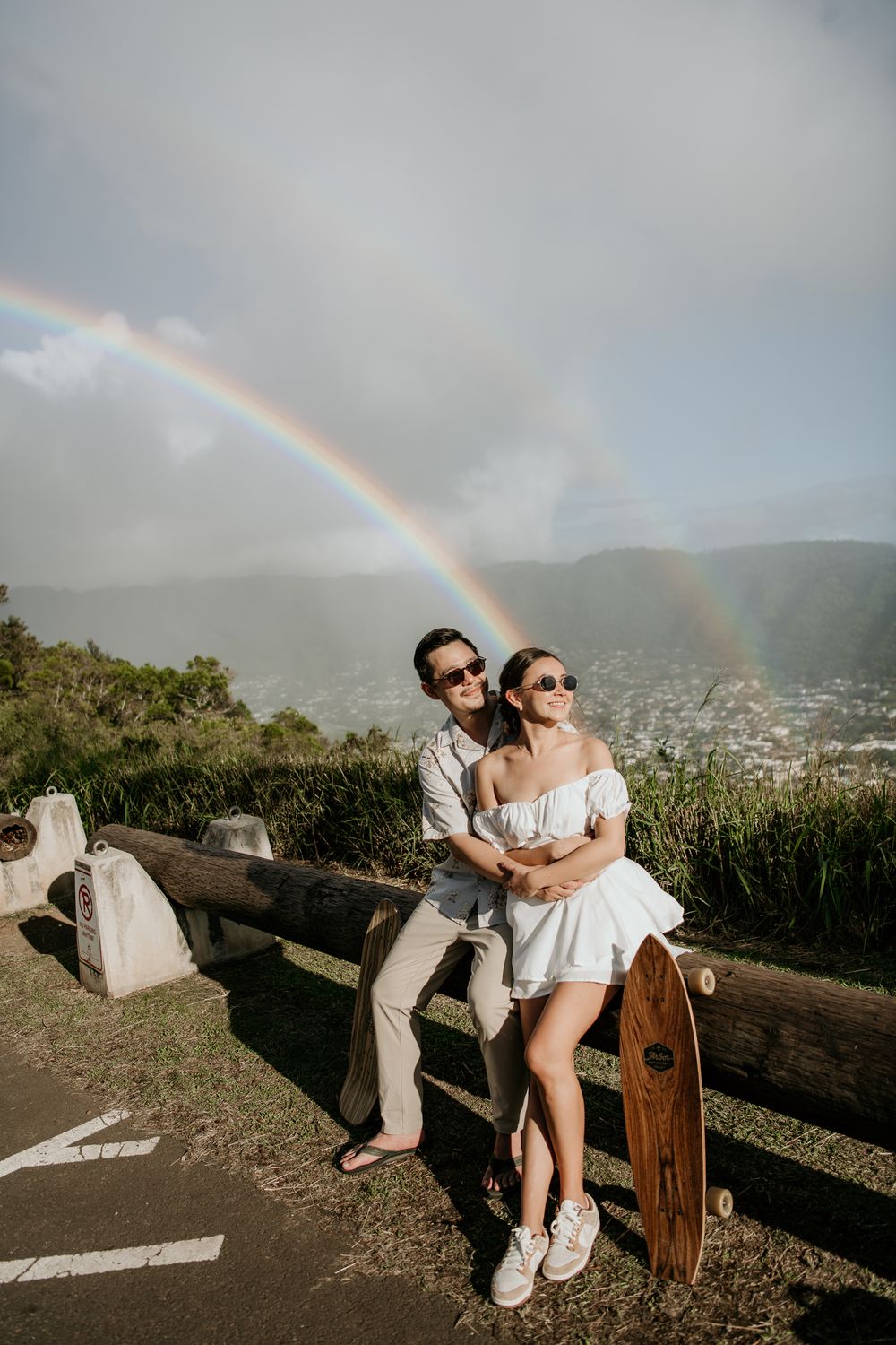A couple leaning against a railing with longboard and double rainbow visible over mountain landscape in Hawaii.