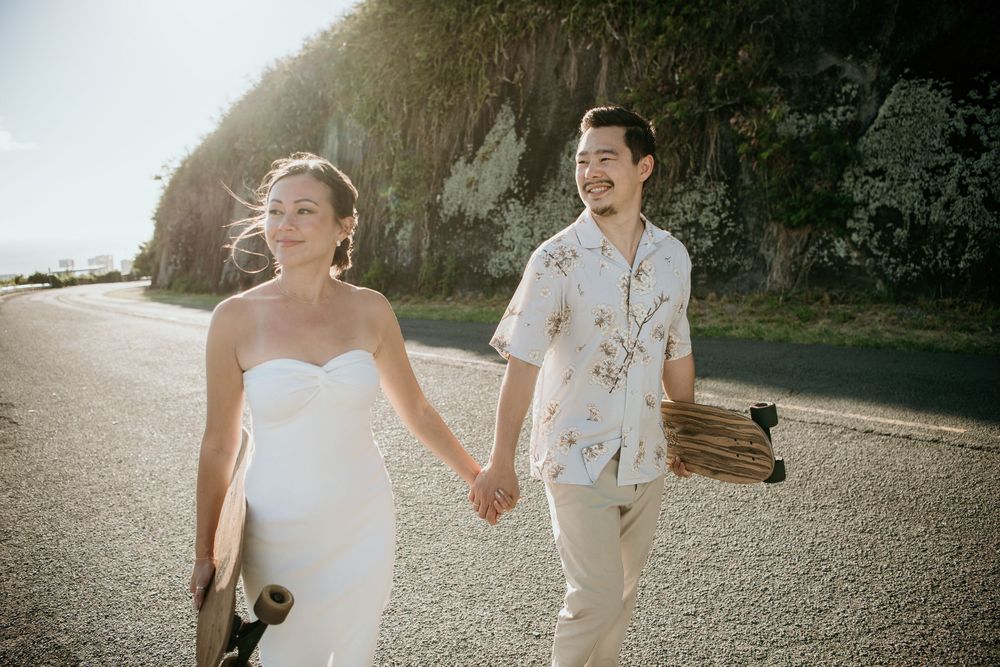 A couple in wedding attire walks hand in hand on a beach path during sunset with cliffs in the background.