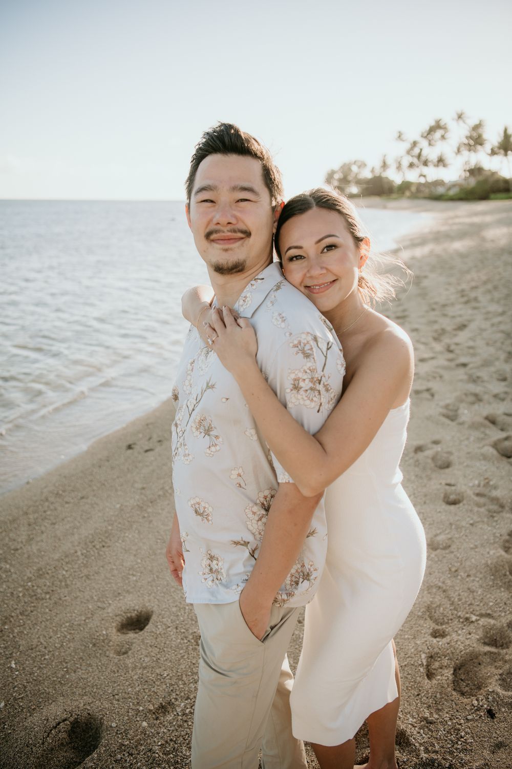 A happy couple embraces on a sunny beach with palm trees and ocean in the background.