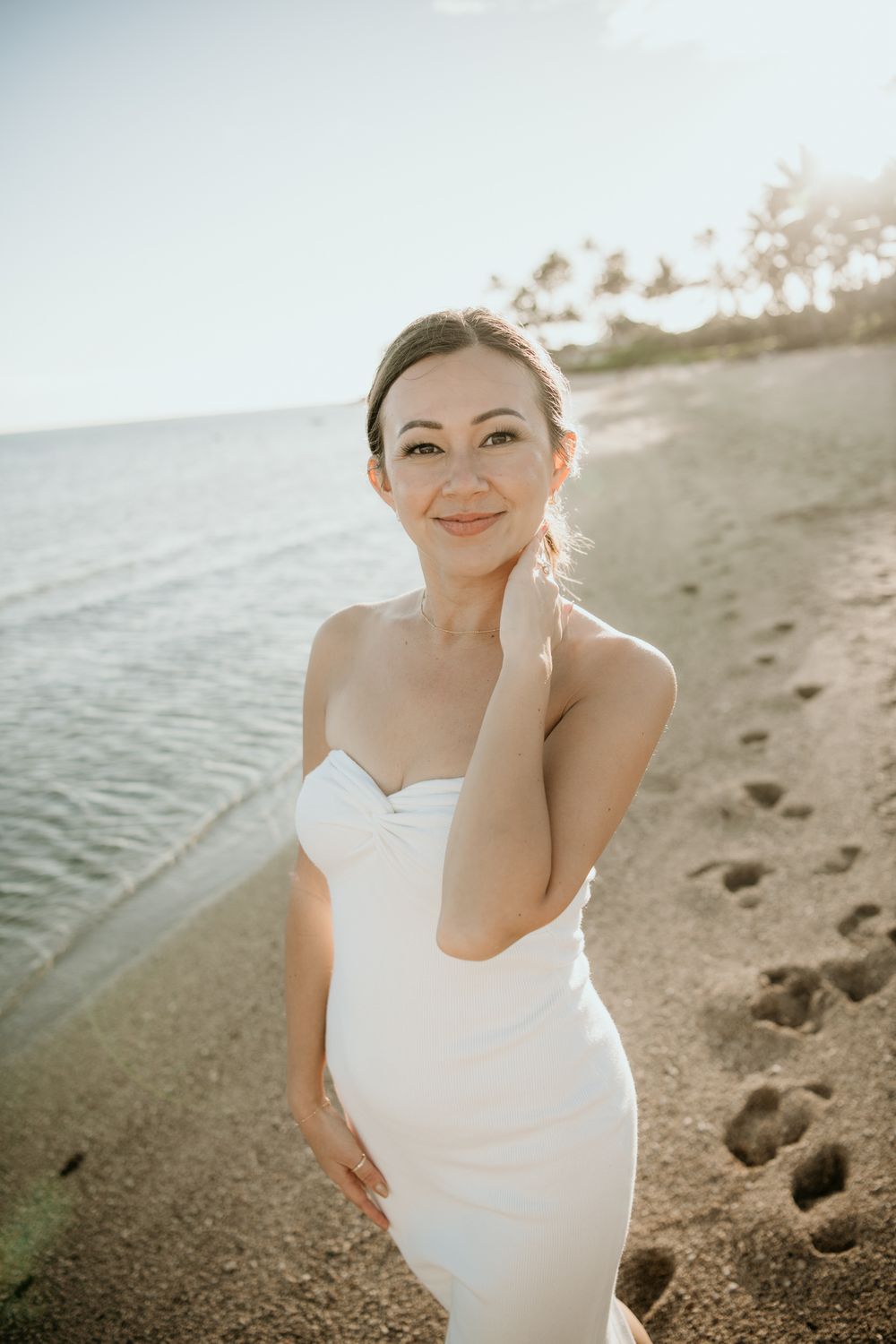 A person in a white dress poses on a beach at sunset with gentle waves and footprints in the sand.