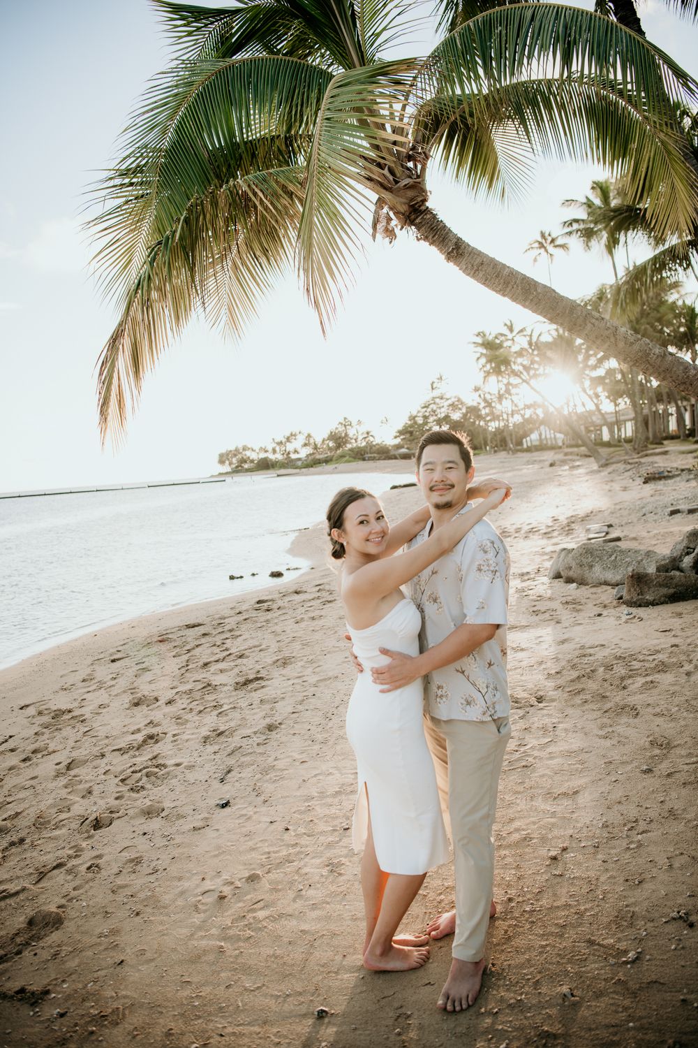 A romantic couple embraces on a tropical beach at sunset with palm trees swaying overhead.