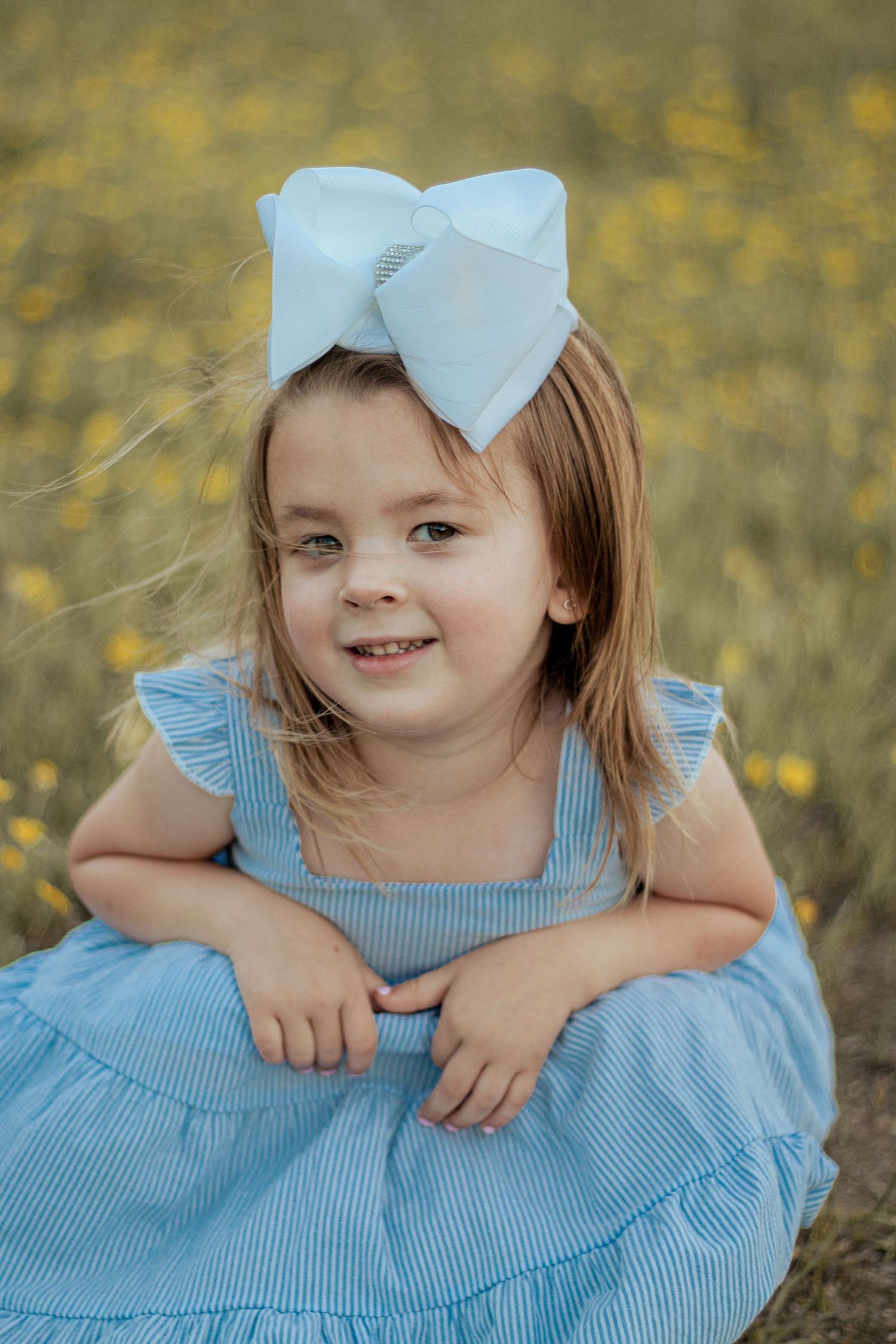 A child in a blue ruffled dress and white bow sits outdoors against a golden bokeh background.