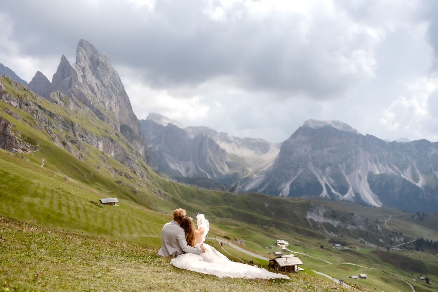 Two people relax on a grassy mountainside with dramatic Dolomite peaks rising in the background under cloudy skies.