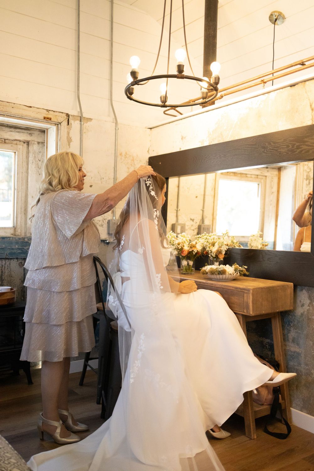 A wedding dress hangs in preparation as someone arranges details in a rustic room with a circular chandelier sussex county nj.