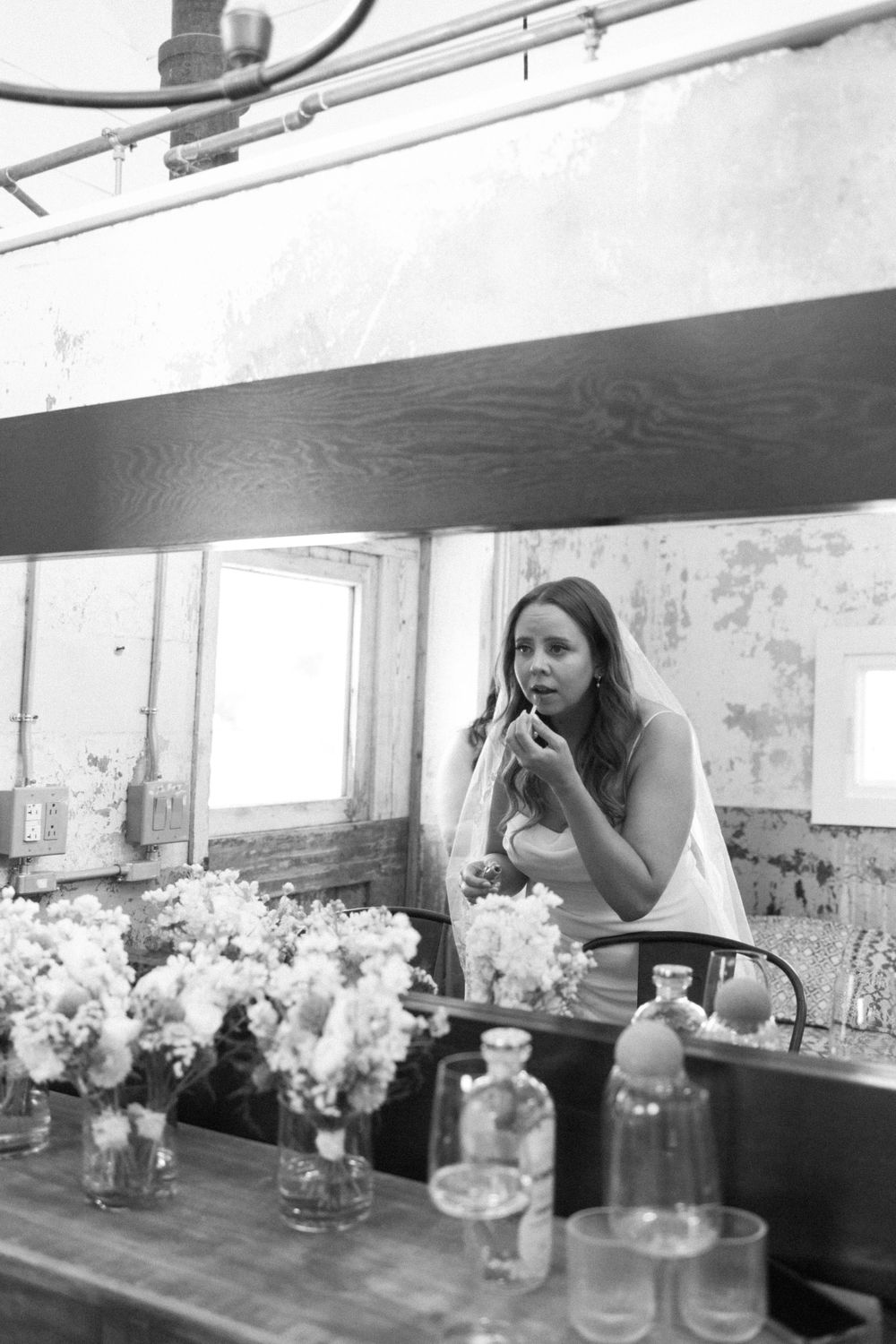 A black and white image shows someone applying makeup at a vanity with mason jars and floral arrangements on the table near vernon nj.