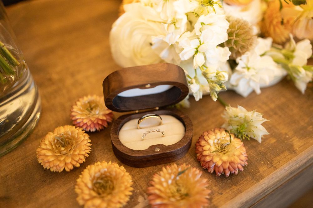 Wedding rings in brown box surrounded by dried orange flowers and white floral arrangements on wooden surface near vernon nj.