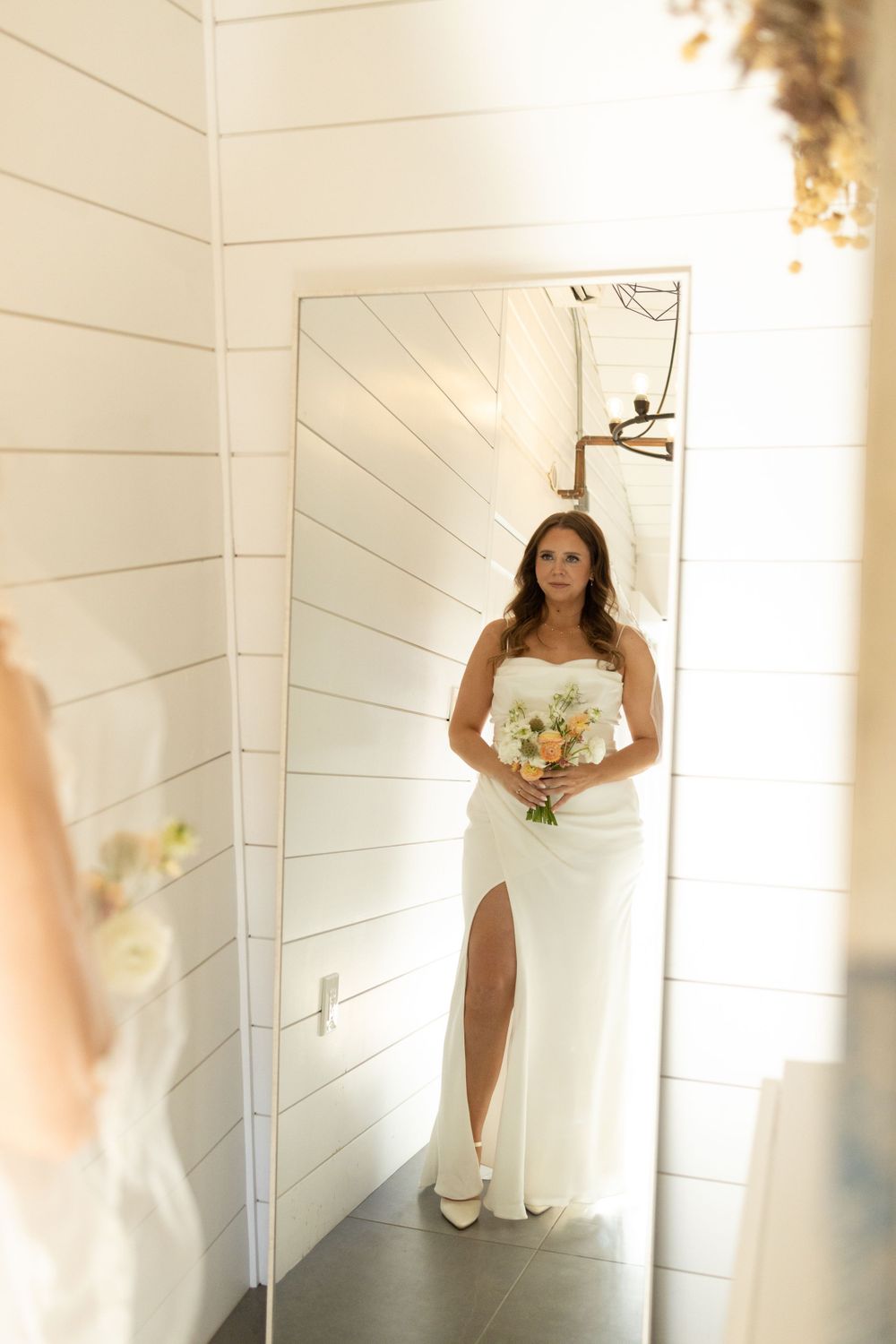 Wedding photo of someone in a sleek white gown with thigh slit holding bouquet, standing in bright corner with shiplap walls near vernon nj.