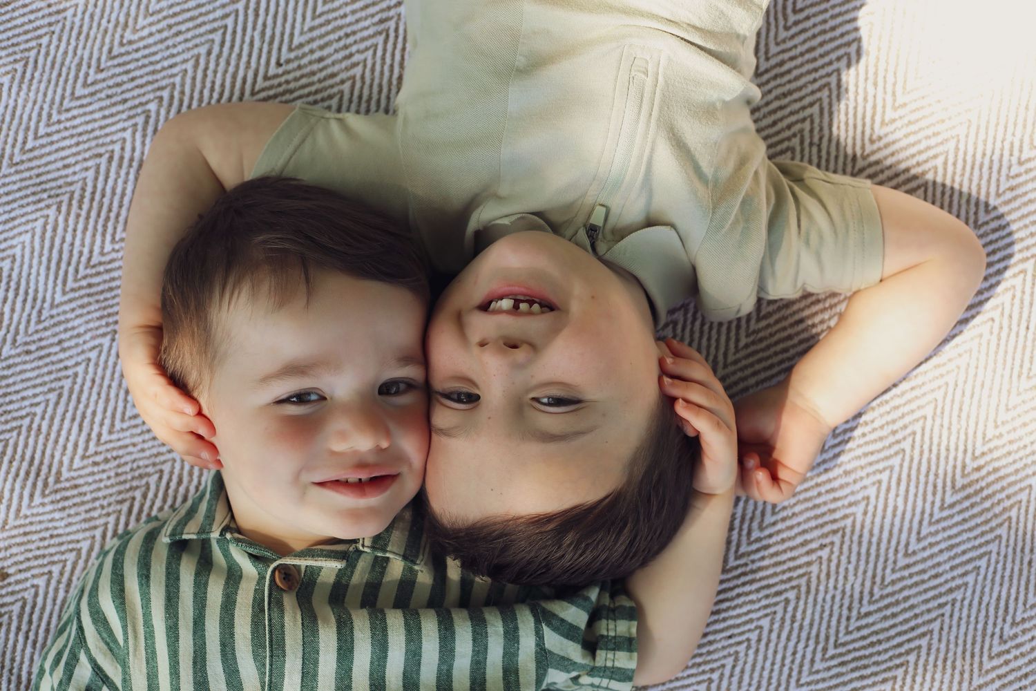 Two children laying on a gray blanket with heads close together smiling at the camera in opposite directions.