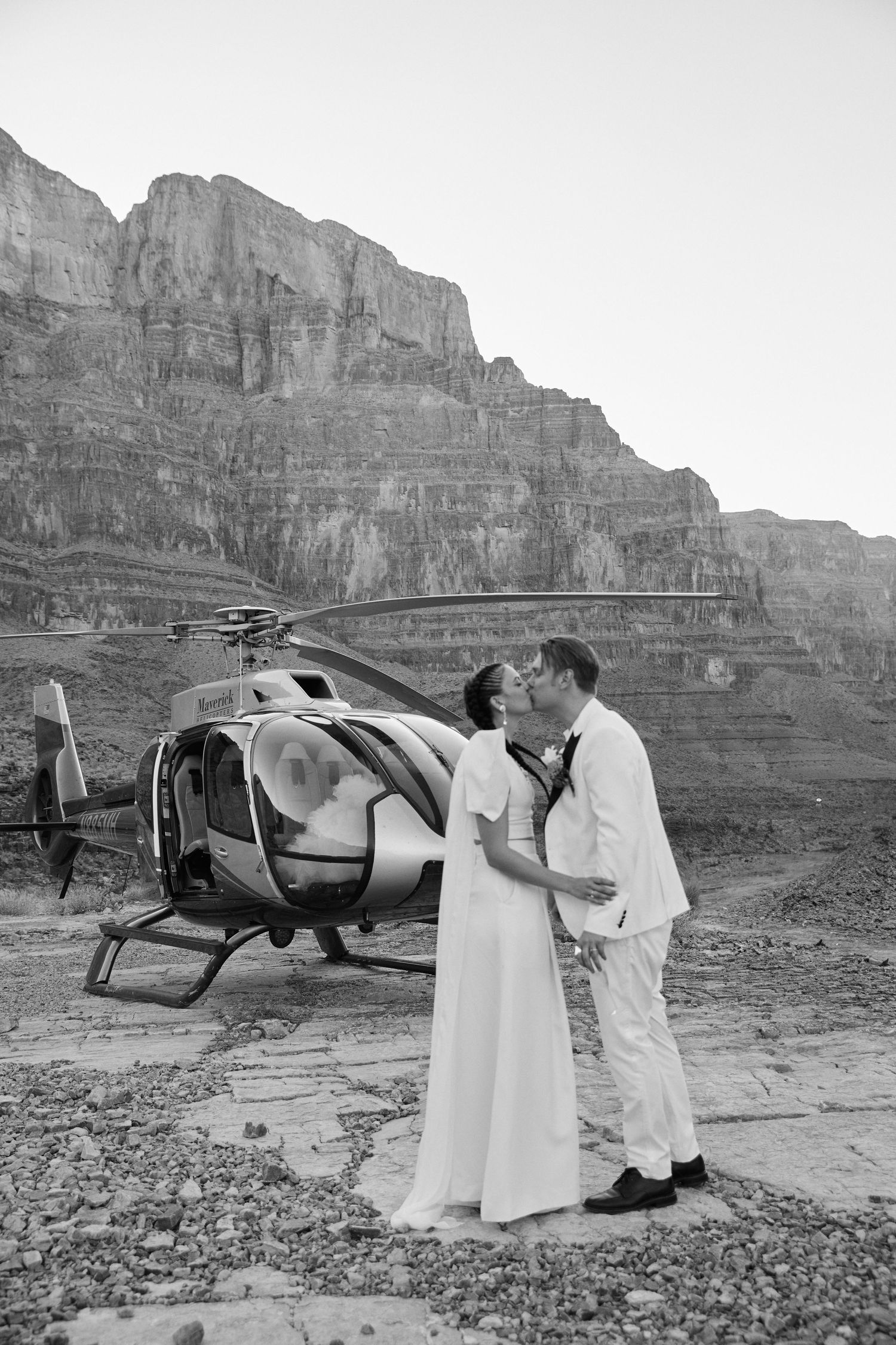 Black and white photo of couple in white outfits posing near helicopter in desert landscape.