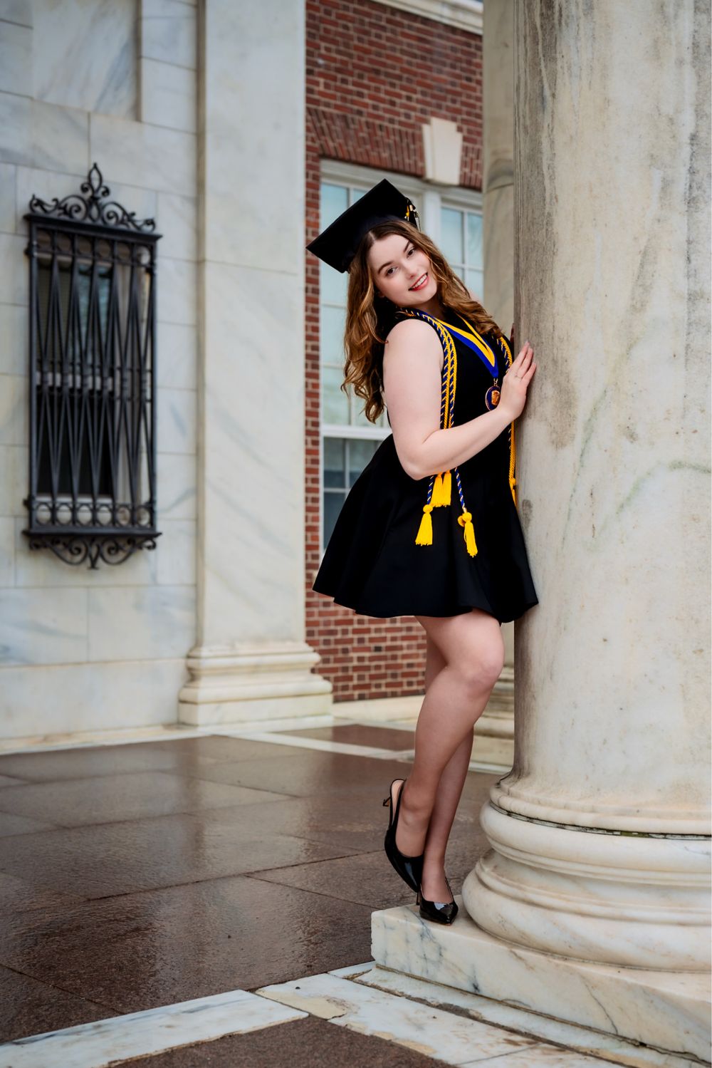 Graduate in black dress and gold stole poses by white column at Davidson Hall.