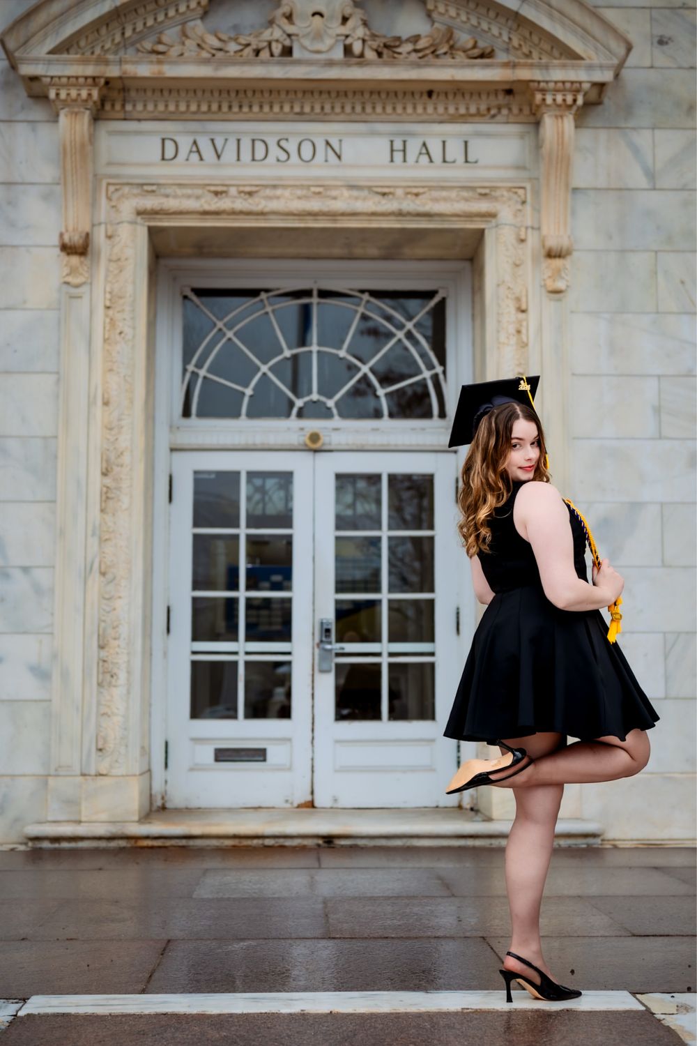 Graduate in black dress celebrates near historic university building entrance.