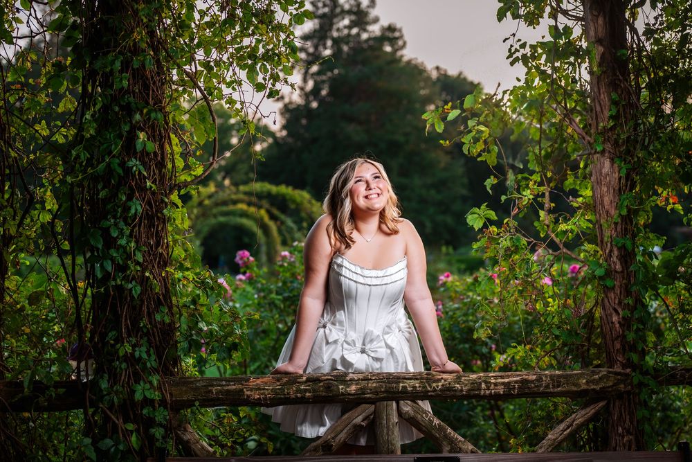 A white dress photoshoot in a lush garden setting with a rustic wooden bridge and greenery-covered archway.