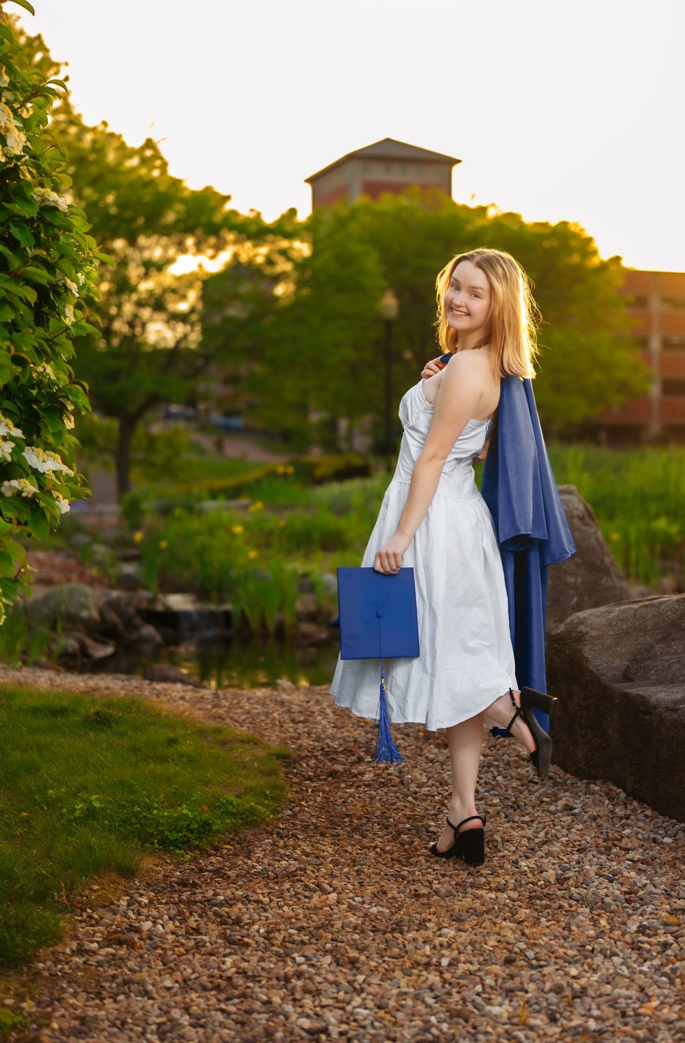 A graduate in a white dress poses with diploma on a rocky path during sunset.