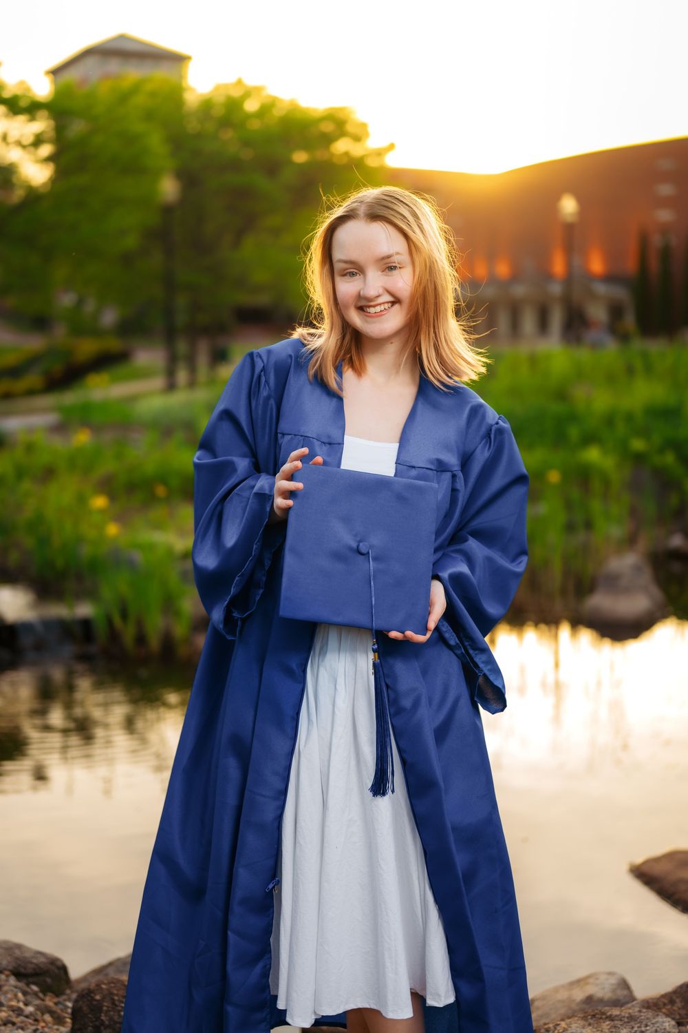 A graduate celebrates with navy blue graduation gown against sunset water backdrop.