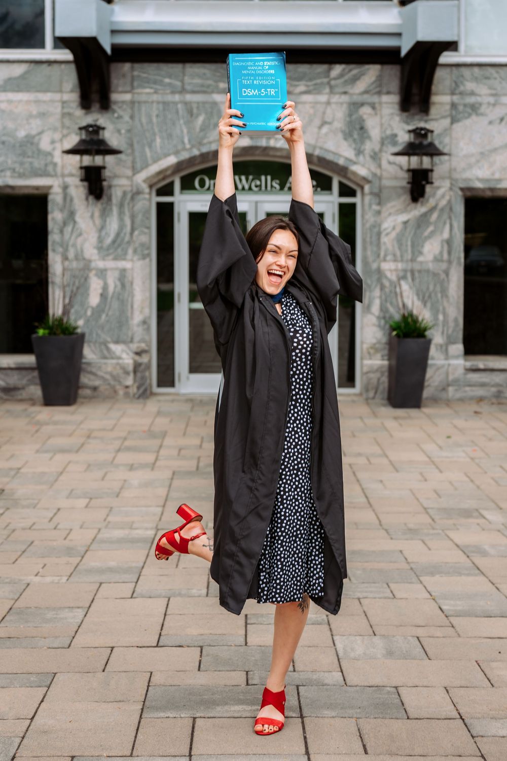 Graduate posing joyfully in black gown and polka dot dress outside stone building.