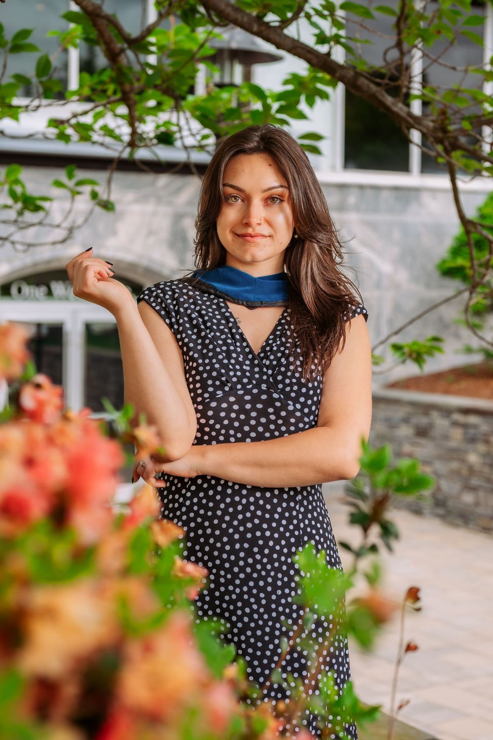 Person in polka dot dress posing playfully near flowering bushes on campus.