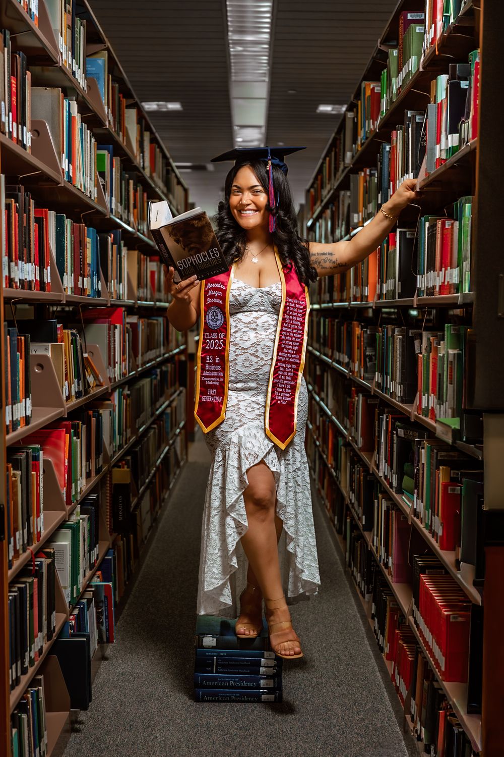 Smiling graduate in cap and gown stole poses between library bookshelves holding a book while standing on stacked books.