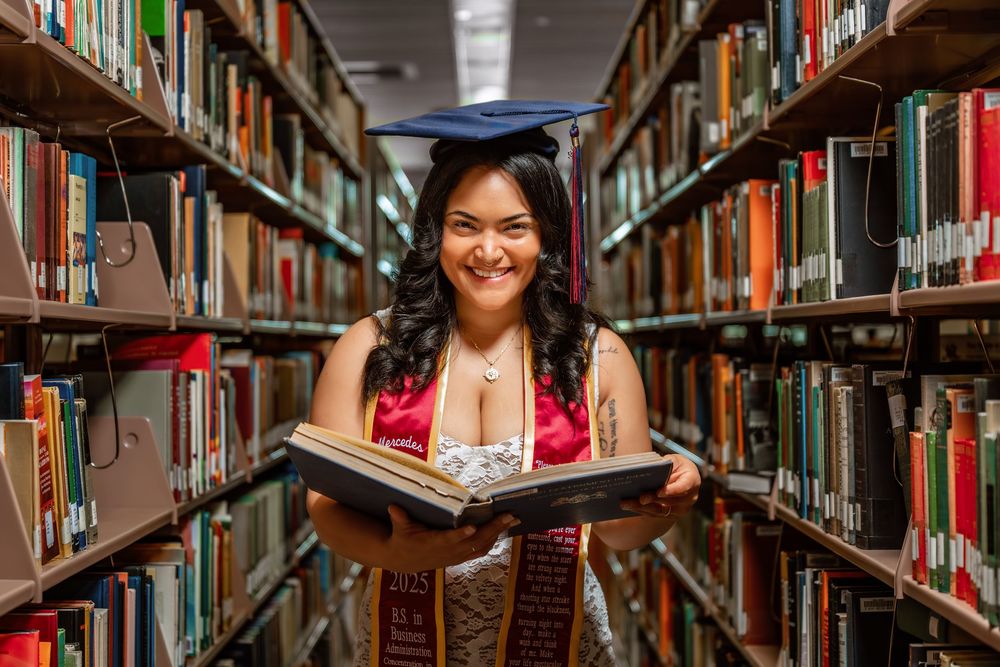Smiling graduate in cap and gown holds open book between library shelves filled with colorful books.