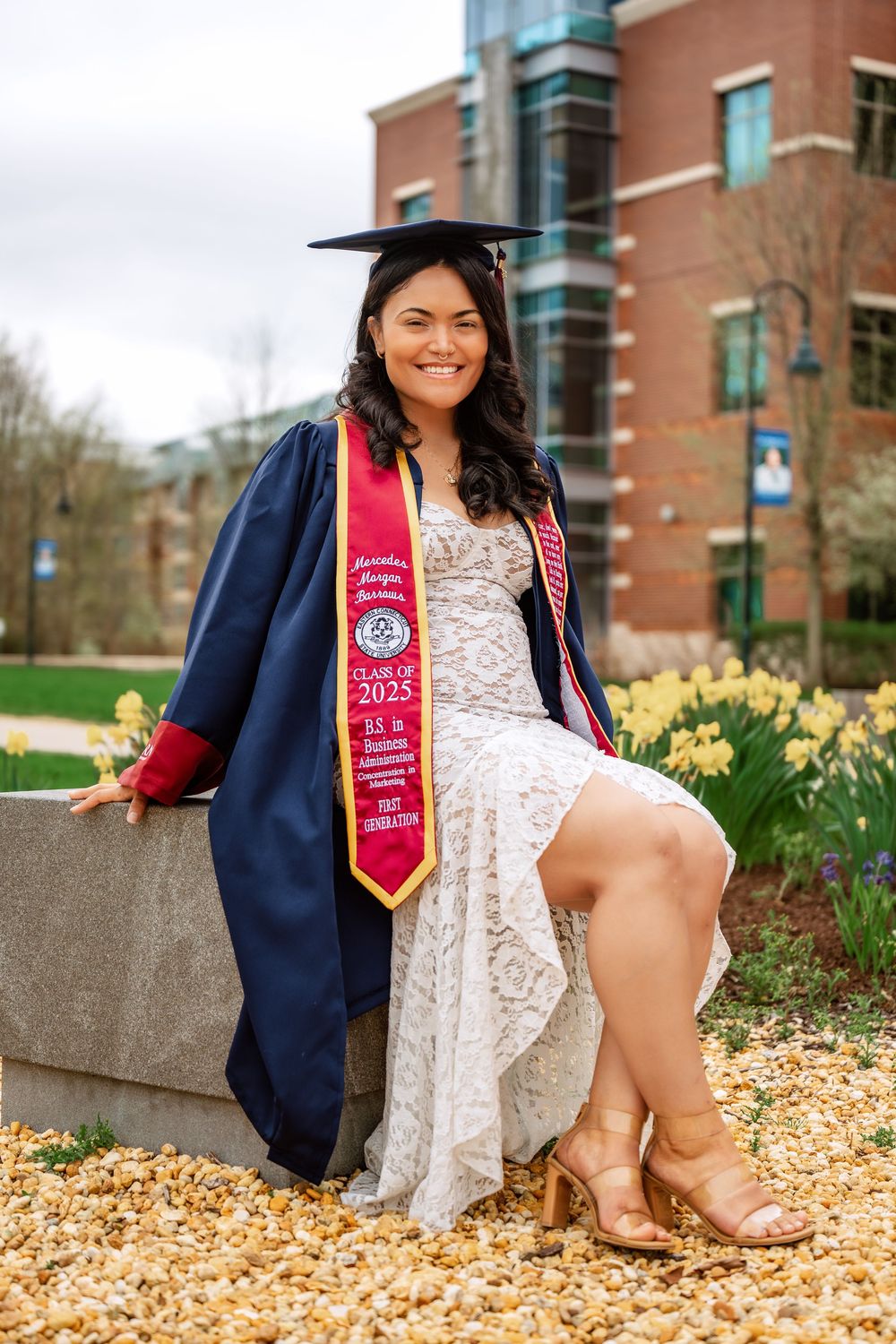 Smiling graduate in navy cap and gown with red stole sits outdoors near yellow flowers on a spring campus.