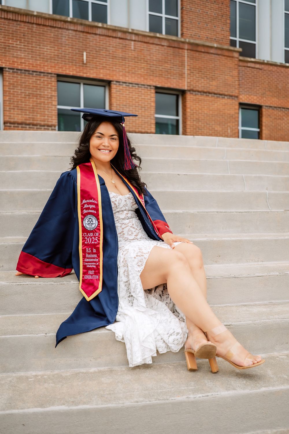 Smiling graduate in navy cap and gown with red stole sits on stone steps outside a brick university building, class of 2025.