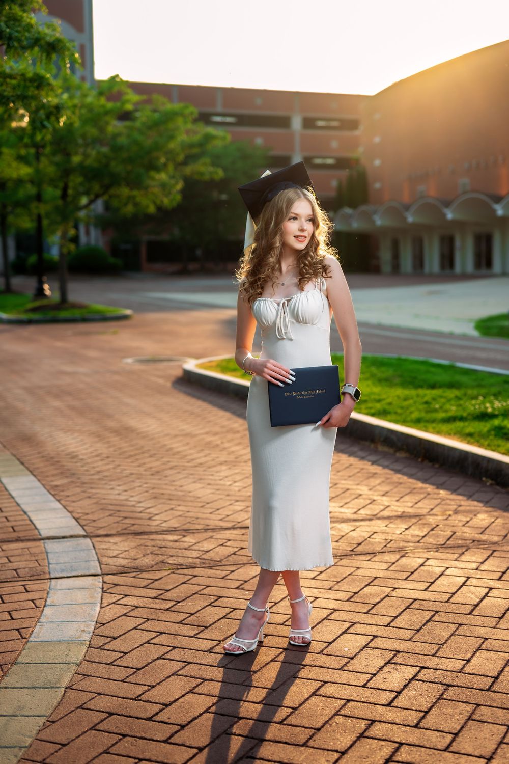 Graduate in white dress poses with diploma on brick pathway at sunset.