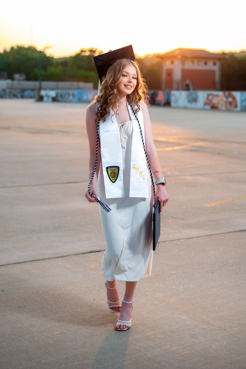 Graduate in white dress walking across campus during golden hour sunset.
