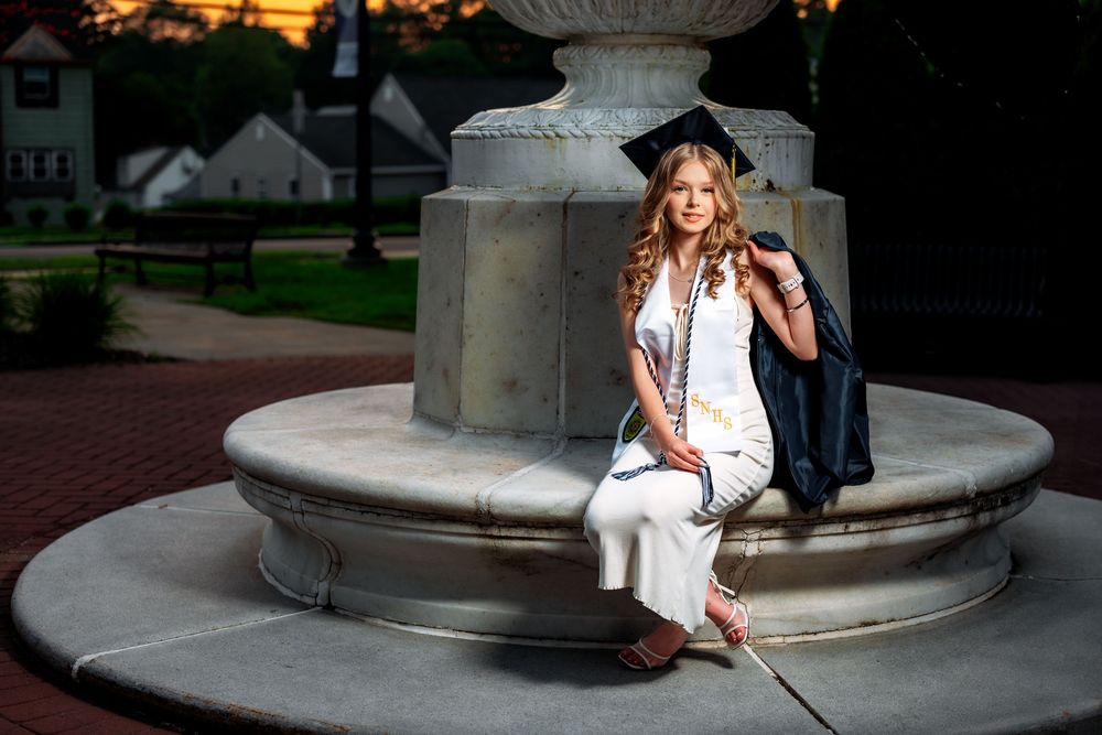 Graduate in white dress sitting on stone fountain at dusk.