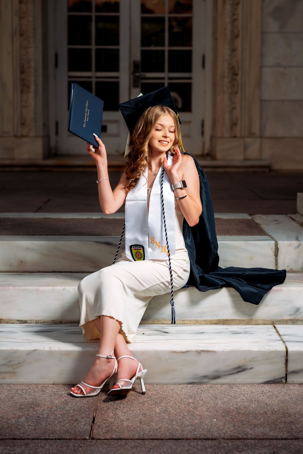 Graduate in white dress posing with diploma on stone steps.