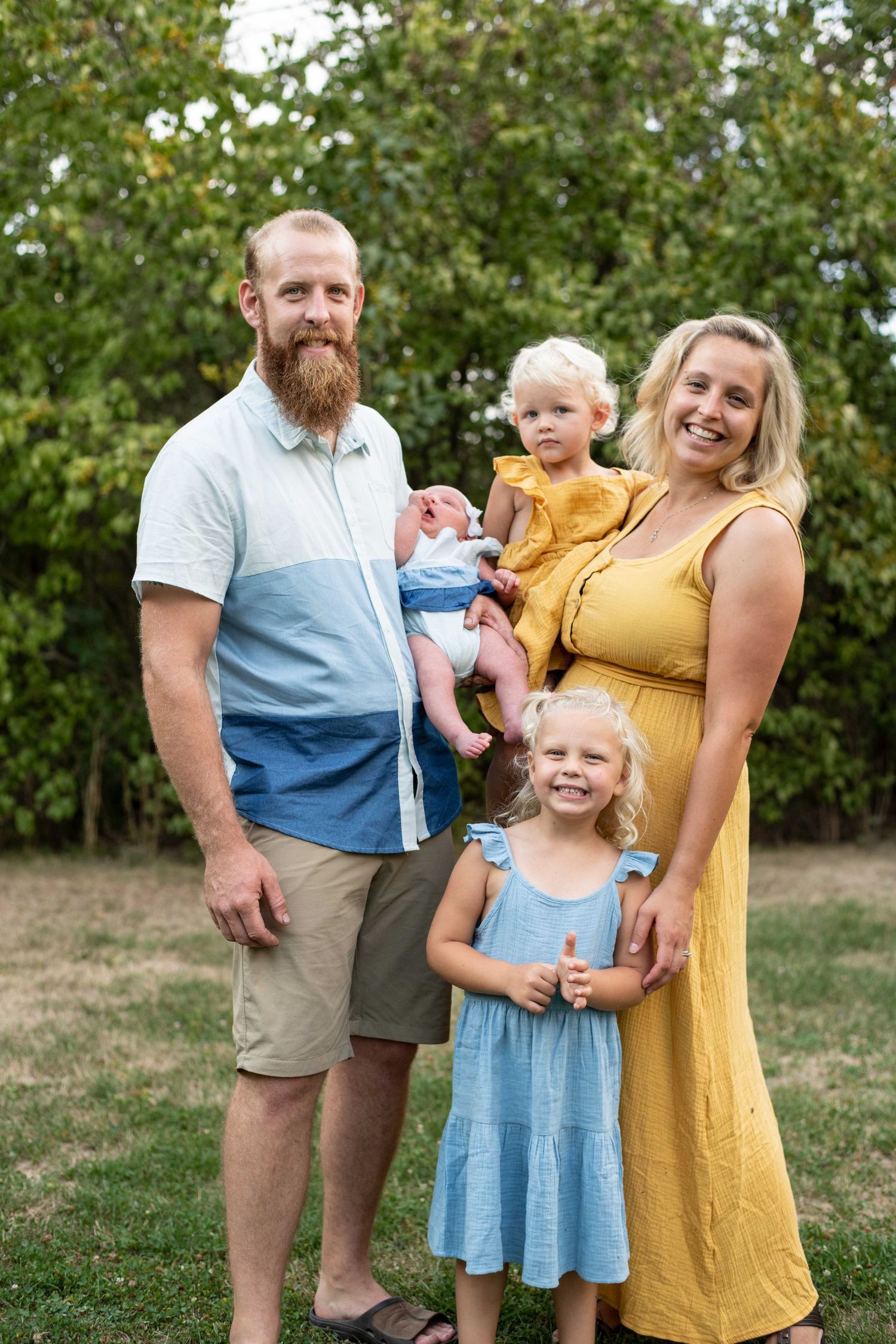 A happy family standing together outdoors on grass with green trees in the background on a sunny summer day.