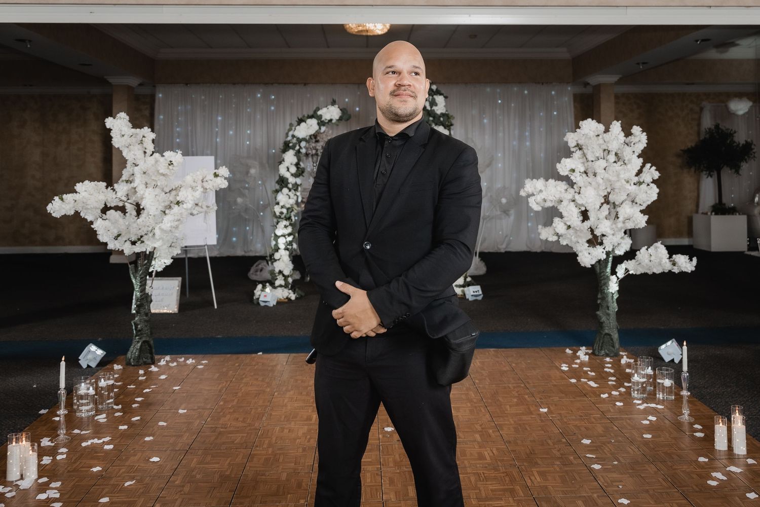 Someone in all black attire stands between floral arrangements and candles in an event space setting.