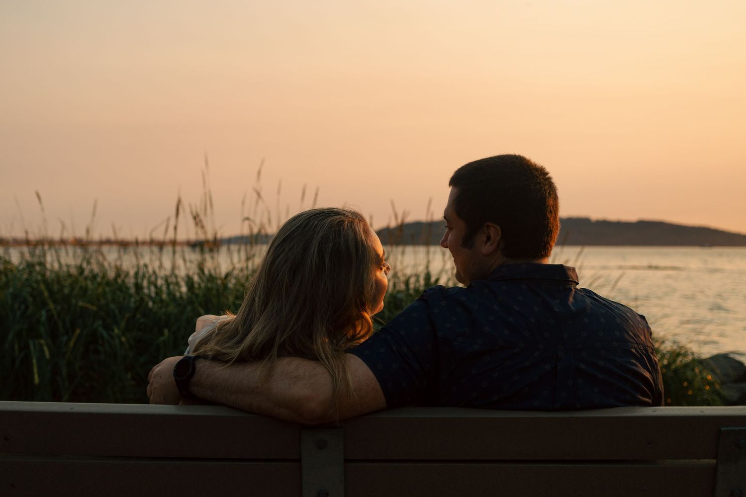 A couple sits together on a bench watching the sunset over a peaceful waterfront.