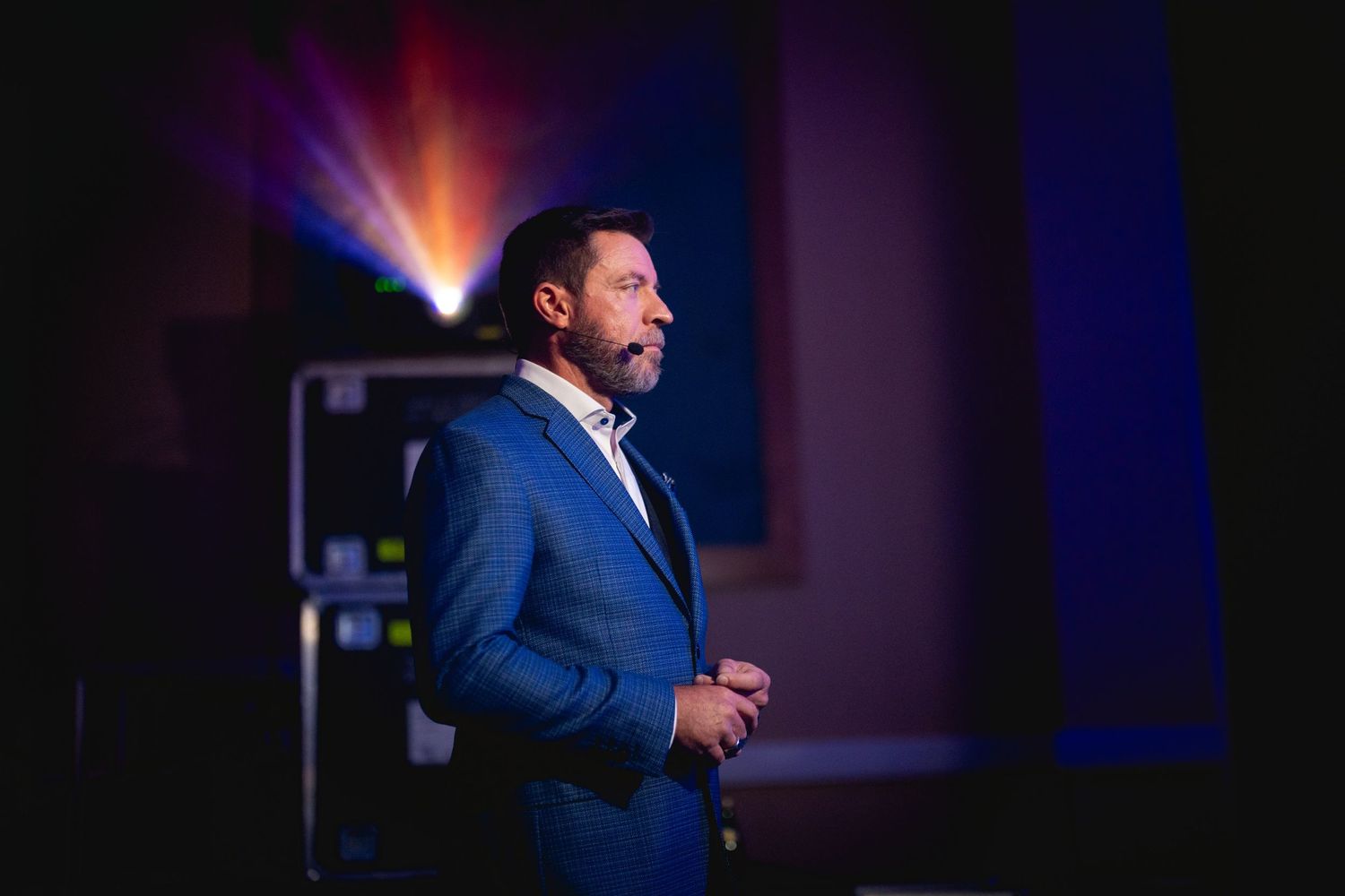Speaker in navy blue suit stands on stage with dramatic colorful lighting in background during presentation.